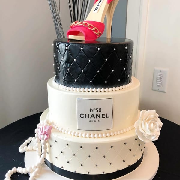A three-tiered Chanel-themed cake with a pink high heel, pearls, and white roses.