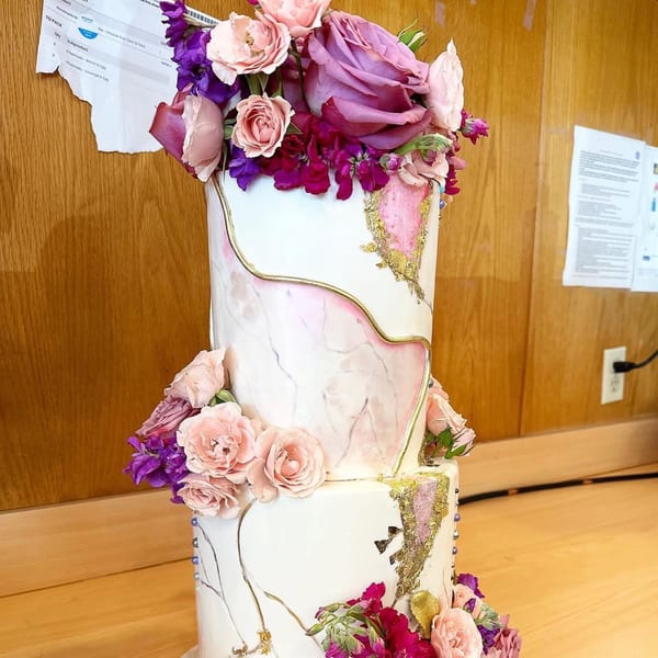 A three-tiered white cake with pink marble, gold accents, and purple and pink floral decorations.