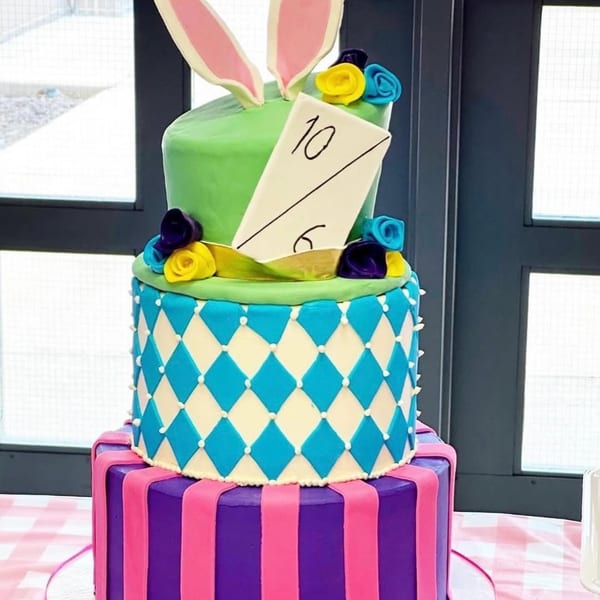 A colorful three-tiered cake with bunny ears, a Mad Hatter card, and harlequin patterns.