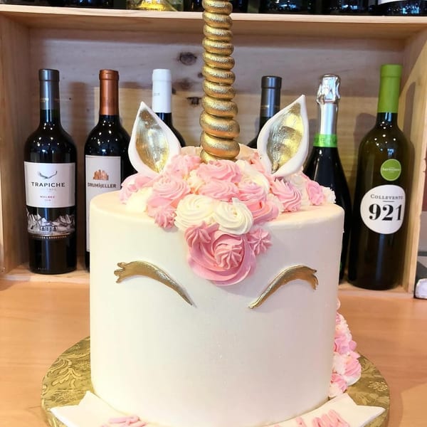A unicorn cake with a golden horn, ears, and eyelashes, decorated with pink and white frosting.