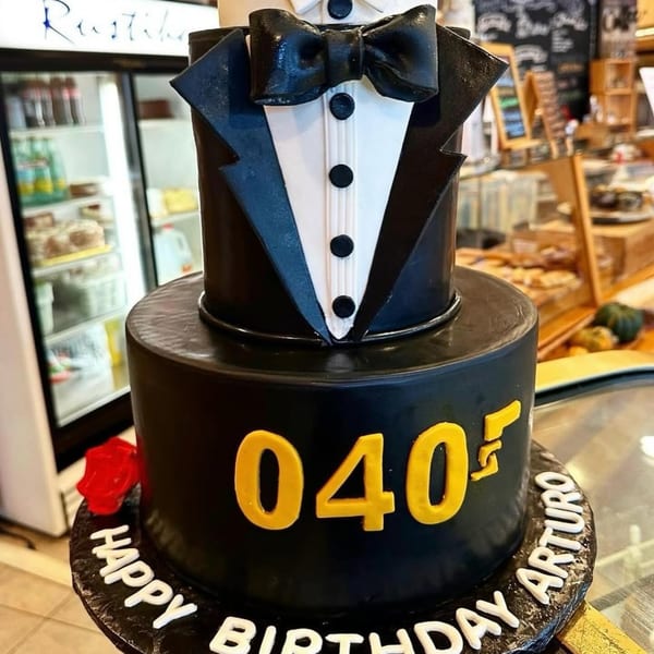 Two-tiered black tuxedo cake with "040" and "Happy Birthday Arturo" in a bakery setting.
