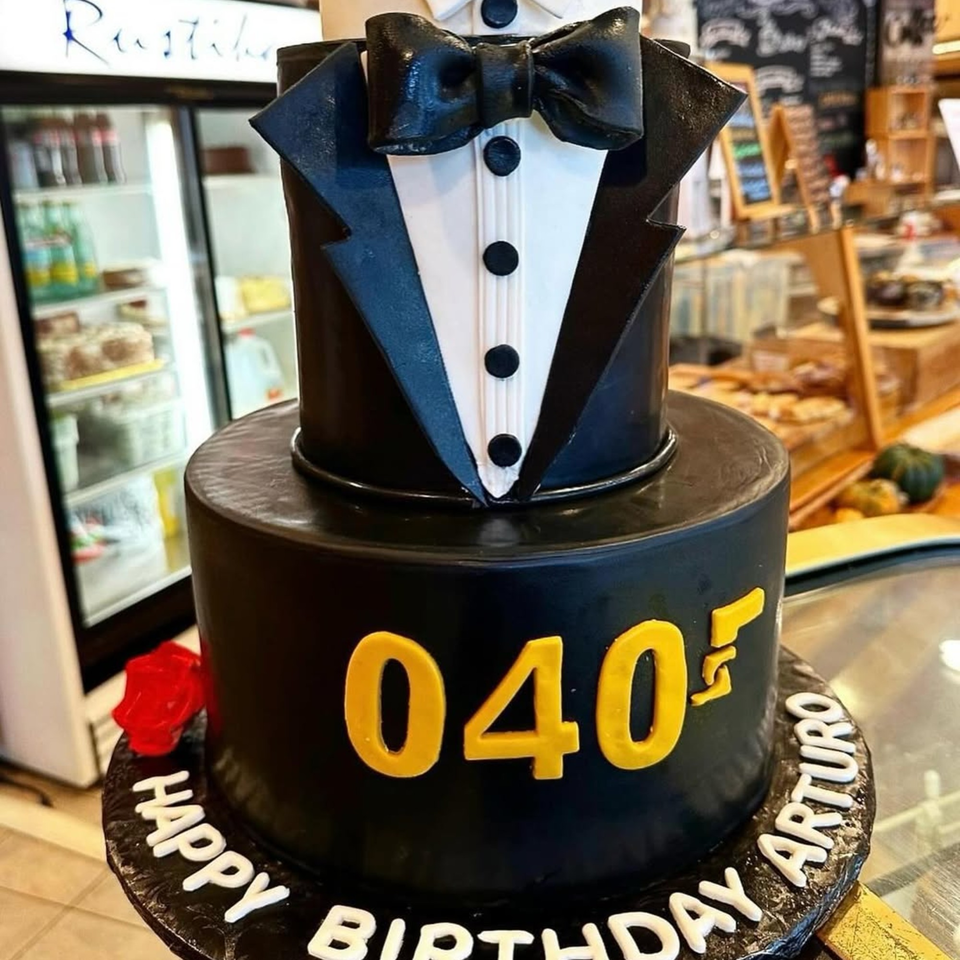 Two-tiered black tuxedo cake with "040" and "Happy Birthday Arturo" in a bakery setting.
