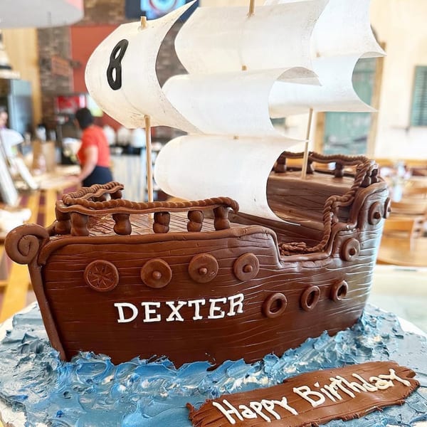 A chocolate pirate ship cake with white sails, blue icing waves, and "DEXTER" written on its side.