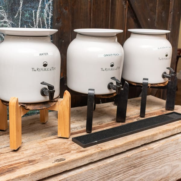 Three white beverage dispensers labeled "Water," "Ginger Peach," and "Blackberry Sage" on a wooden counter.