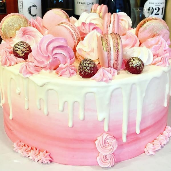 Pink ombre cake with white drip icing, topped with macarons, meringues, and gold-dusted raspberries.