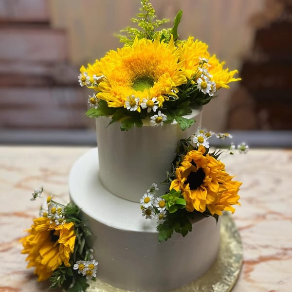 Two-tiered white cake adorned with sunflowers, daisies, and greenery, perfect for Mama's Bistro.