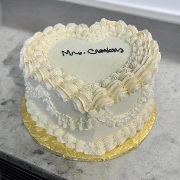 A heart-shaped white cake with "Mrs. Crowlens" written in black icing, decorated with white frosting swirls.