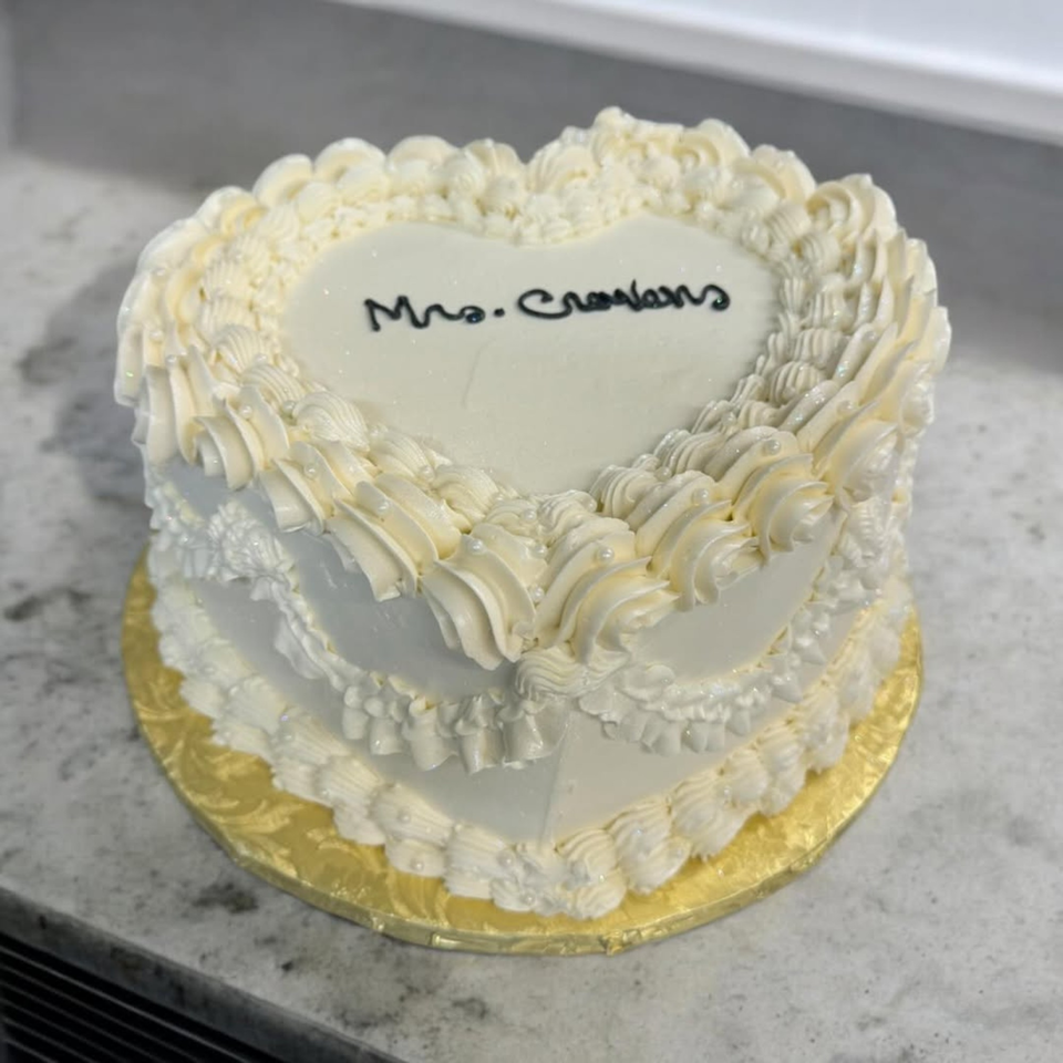 A heart-shaped white cake with "Mrs. Crowlens" written in black icing, decorated with white frosting swirls.