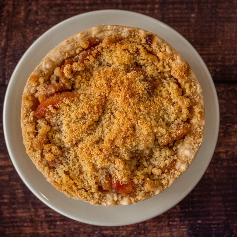 Overhead view of a golden-brown apple crumble pie on a light plate, on a dark wooden table.