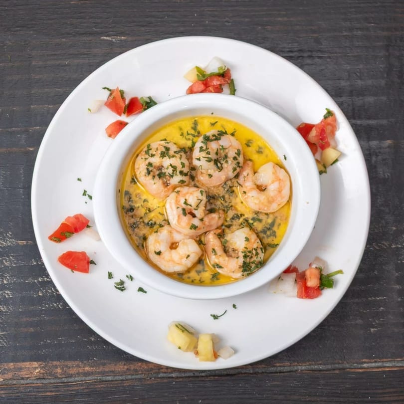 Shrimp scampi in a white bowl with a yellow sauce and parsley, garnished with diced tomatoes.