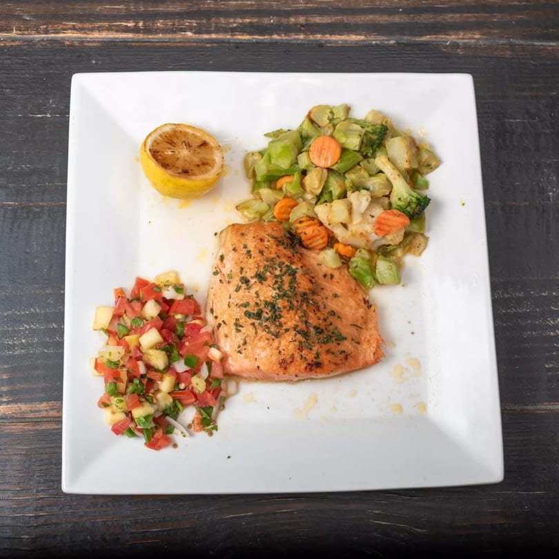 Grilled salmon with roasted vegetables, fresh salsa, and a lemon half on a white plate.