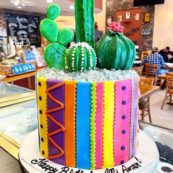 A vibrant, multi-colored cake with cactus decorations and "Happy Birthday Mi Amor!" written on the base.