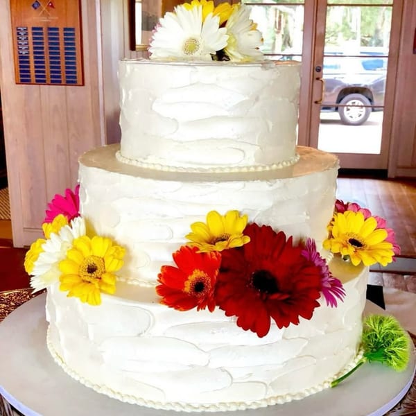 Three-tiered white cake with colorful daisies, including red, yellow, white, and pink, on a table.