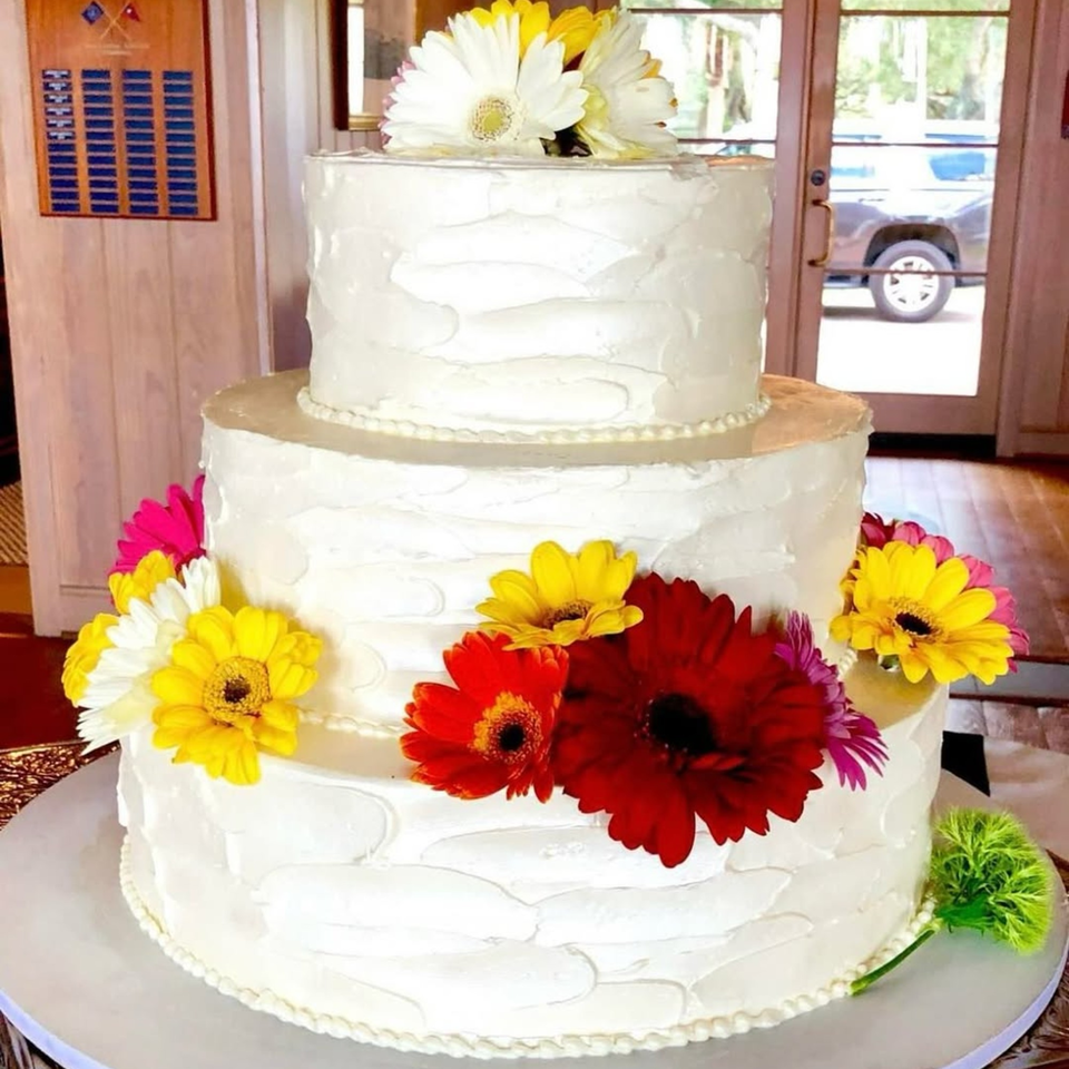 Three-tiered white cake with colorful daisies, including red, yellow, white, and pink, on a table.