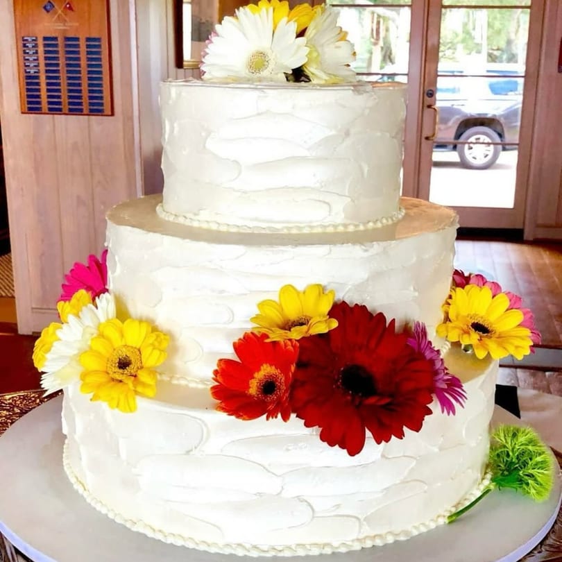 Three-tiered white cake with colorful daisies, including red, yellow, white, and pink, on a table.